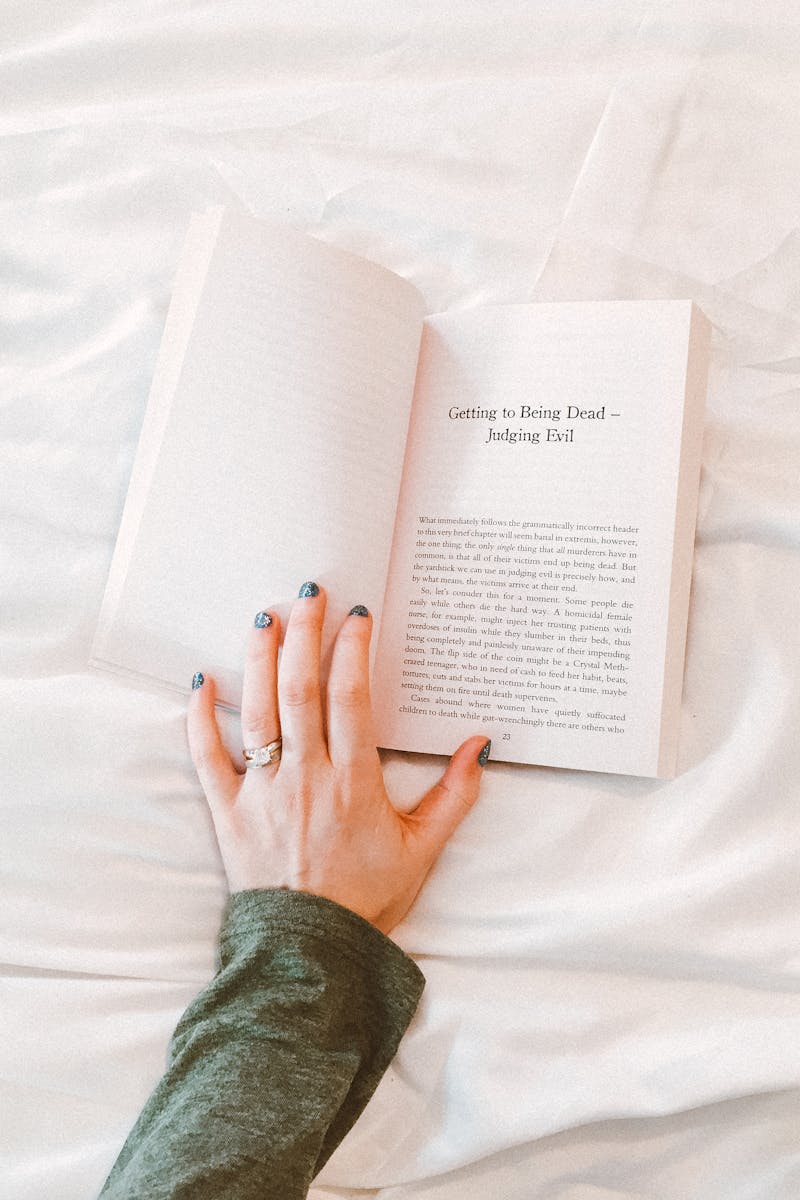 Close-up of a woman's hand holding a book while relaxing on a bed with white sheets. Perfect for lifestyle themes.