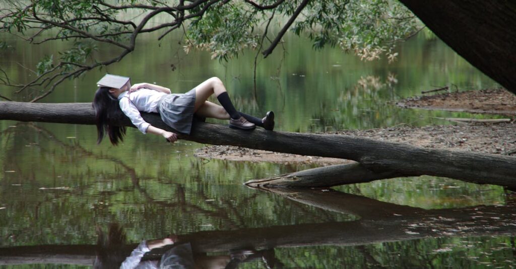 A young woman with a book on her face lies relaxing on a tree trunk by a tranquil lake.