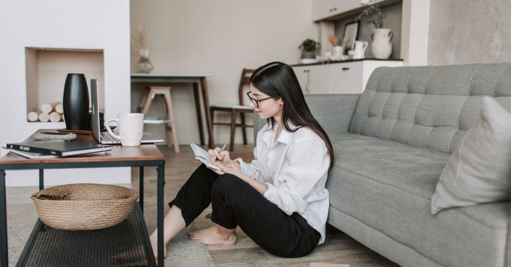 Side view of thoughtful young woman in eyeglasses and casual outfit sitting on floor near sofa and writing in notebook while resting in comfortable apartment