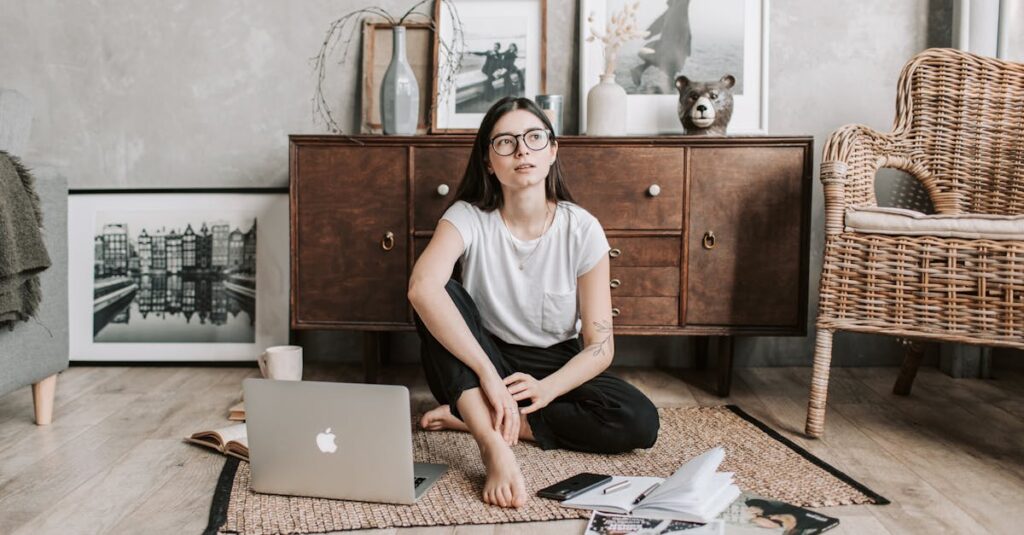 A calm woman with eyeglasses sitting barefoot at home, contemplating and relaxing with a laptop and magazines.