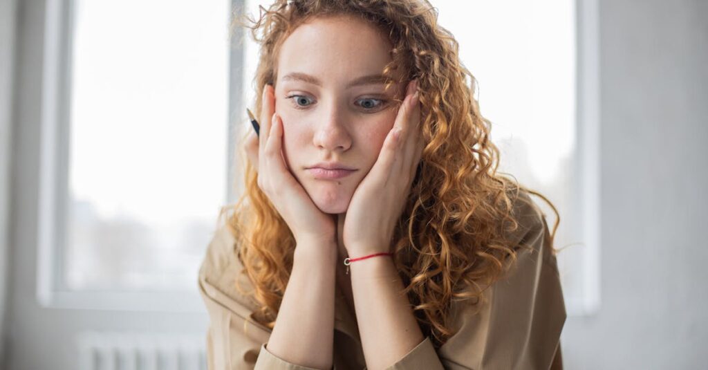 Pensive female student with hands on cheeks looking down while sitting in light room on blurred background while deciding task in frustration